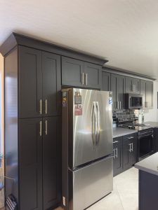 Iron black shaker kitchen cabinets with crown molding and a tall pantry beside a stainless steel refrigerator. 